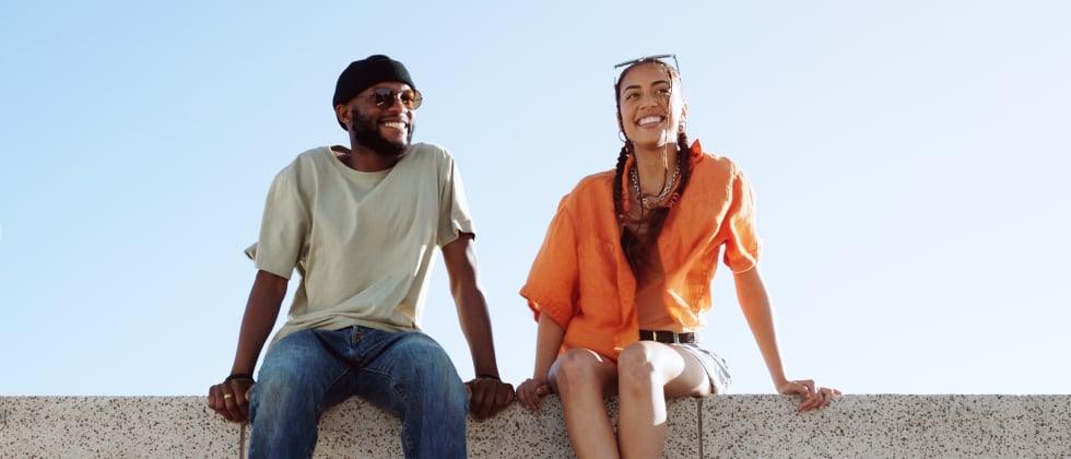 A woman and a man sitting next to each other outside. They are both smiling and their hands are slightly touching.