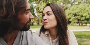 A man and a woman sitting next to each other in nature. She is looking at him happily and showing a love language.