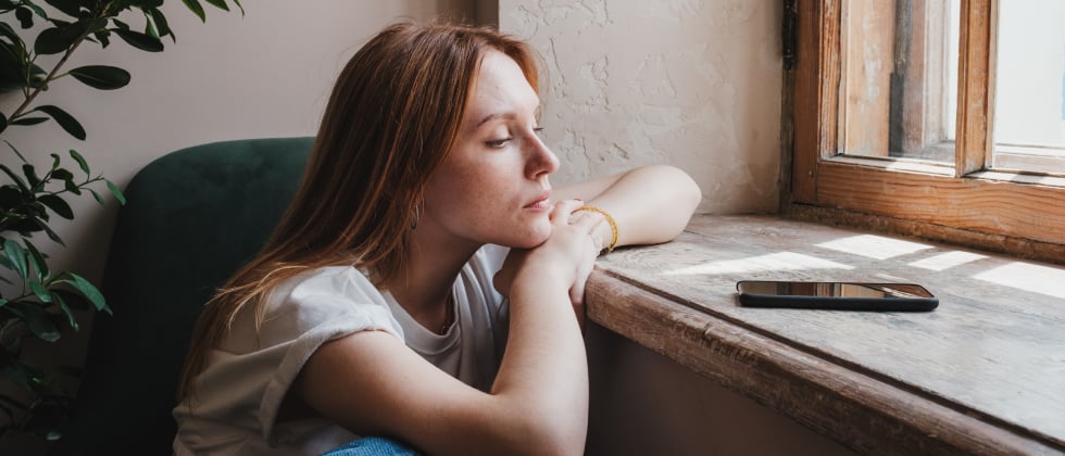 A woman experiencing unrequited love, looking at her phone while sitting at a window.